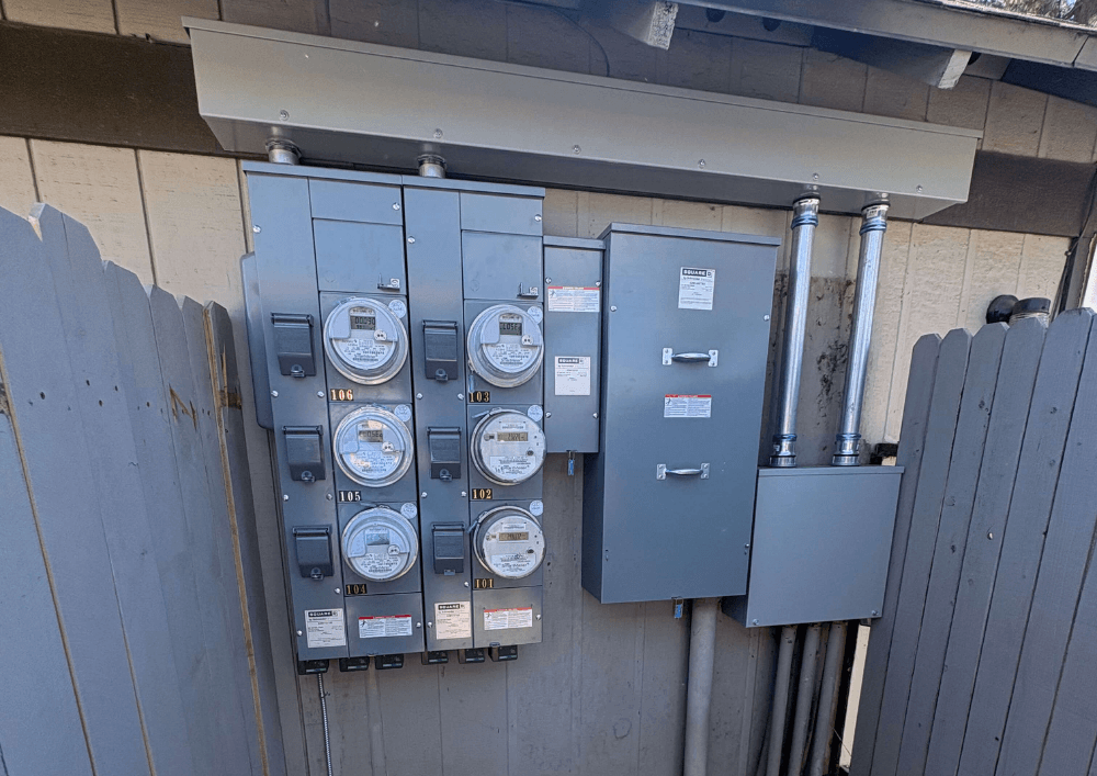 Electrical meter panels and utility boxes mounted on an exterior wall behind a gray wooden fence, with six electric meters and a paved stone walkway below.